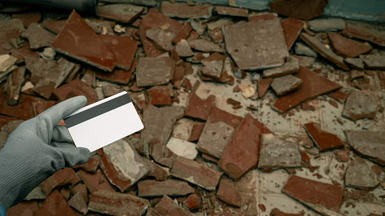 woman's hand adorned in construction gloves, holding a bank card, set against a backdrop of shattered tiles on the floor. Symbolizing the initiation of renovation and the considerable expenses associated with repairs.