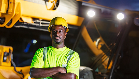 A construction worker standing in front of a telescopic handler. It is early morning, still dark out. The worker is a mid adult African-American man wearing a hardhat and reflective clothing.