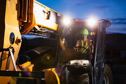 A construction worker operating a telescopic handler. It is early morning, still dark out. The worker is a young Hispanic woman wearing a hardhat and safety vest.