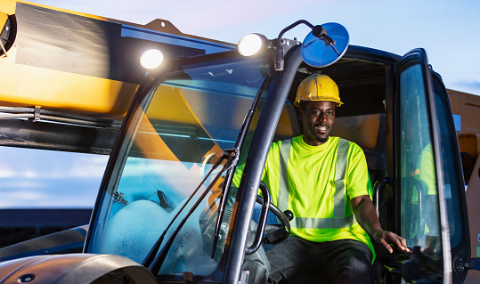 A construction worker operating a telescopic handler. It is early morning. The worker is a mid adult African-American man wearing a hardhat and reflective clothing. He is sitting in the driver's seat, looking out the open door, toward the camera.