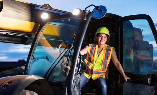 A construction worker operating a telescopic handler. It is early morning. The worker is a young Hispanic woman wearing a hardhat and safety vest. She is looking out the open door, smiling.