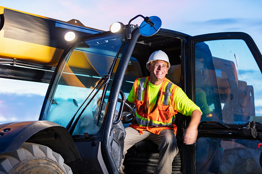 A construction worker operating a telescopic handler. It is early morning. The worker is a mature man wearing a hardhat and safety vest.
