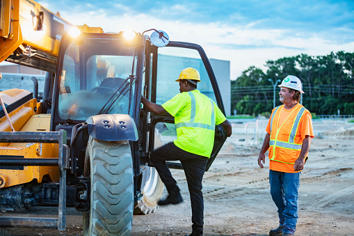 Two multiracial construction workers working at a job site. The African-American man, in his 30s, is climbing into a construction vehicle, a telescopic handler. A foreperson, a mature man in his 50s, is watching.