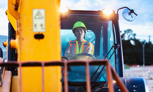 A construction worker operating a telescopic handler. It is early morning. The worker is a young Hispanic woman wearing a hardhat and safety vest.