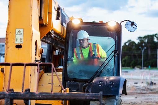 A construction worker operating a telescopic handler. The worker is a mature man wearing a hardhat and safety vest.