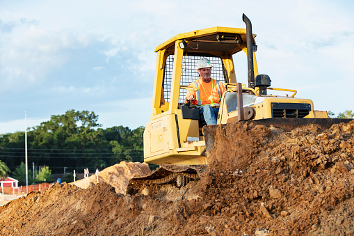 A construction worker operating a bulldozer. The worker is a mature man wearing a hardhat and safety vest.
