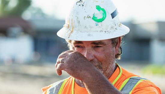 Face of a serious construction worker wearing a hardhat and reflective vest. He is looking down, thinking, wiping his face with his hand.