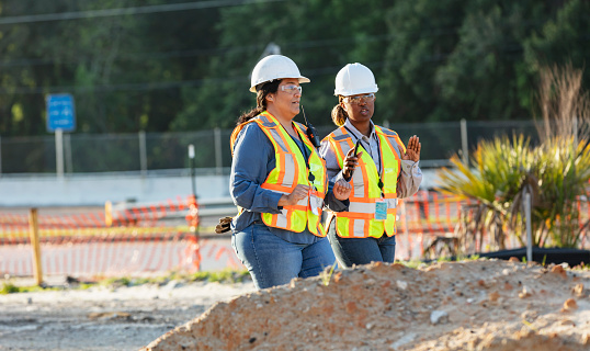 Two mature multiracial women working as construction workers or engineers, walking side by side through a job site, conversing. They are wearing hard hats, reflective vests and safety glasses. Piles of dirt are in the foreground and a road is in the background.