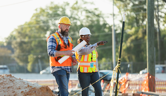 Two multiracial construction workers or engineers walking side by side through a construction site, conversing. One of them is a mature African-American woman in her 40s. Piles of dirt are in the foreground and a road is in the background.