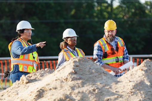 A multiracial group of three construction workers, two women and one man, walking through a construction site, conversing and looking around. Piles of dirt are in the foreground and a road is in the background.