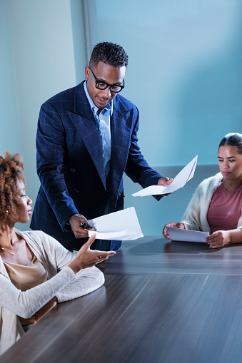 A mid adult African-American businessman standing in a meeting room handing out papers. Two young businesswomen are sitting at the conference table. The focus is on the man, who is about to give a presentation or teach a seminar.