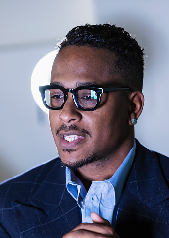 Headshot of an African-American businessman, in his 30s. He is wearing eyeglasses and earrings, looking away from the camera with his hands clasped.