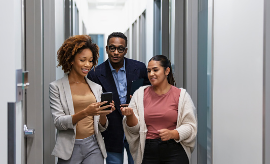 A multiracial group of three office workers walking down an office corridor smiling and conversing. The young African-American woman shows something on her mobile phone to her coworkers.