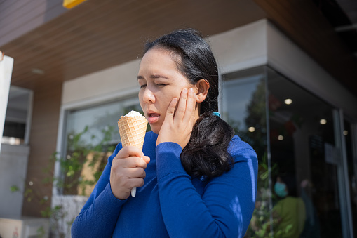Adult Asian female eating ice cream outside cafe and having toothache by observing from her facial expression and using hand to cover the illness.