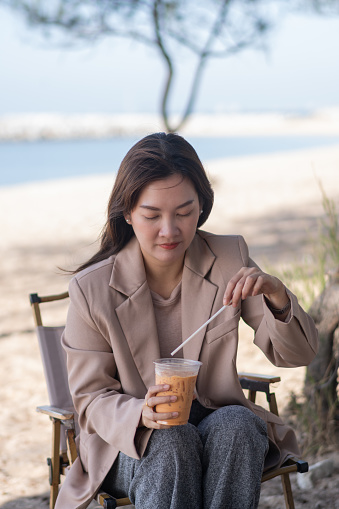 Asian woman with brown formalwear sitting at a cafe at the beach and using straw for drinking the cold beverage for reducing the pain of toothache.