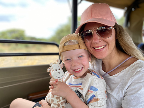 A mother is on safari in Namibia with her 4-year-old son. He is holding a stuffed giraffe in his hand and they are both smiling at the camera