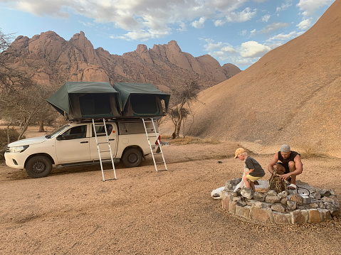 A father and his 4-year-old son build a campfire in the fireplace in Namibia while an off-road vehicle with roof tents is parked in the background