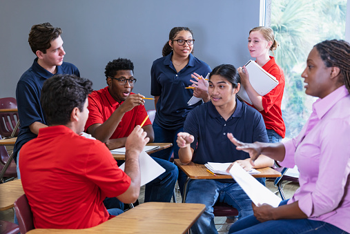 A multiracial group of six high school students sitting together in a classroom having a discussion, with a teacher, an African-American woman. The students are in two teams, wearing either blue or red shirts. They could be participating in an after school activity, perhaps a debate team or math club.