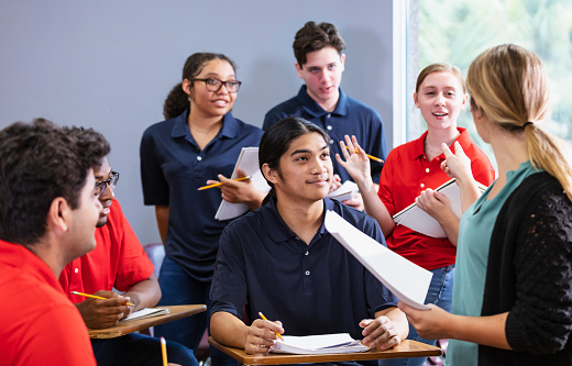 A multiracial group of six high school students sitting together in a classroom having a discussion, with a teacher leading. The students are in two teams, wearing either blue or red shirts. They could be participating in an after school activity, perhaps a debate team or math club.