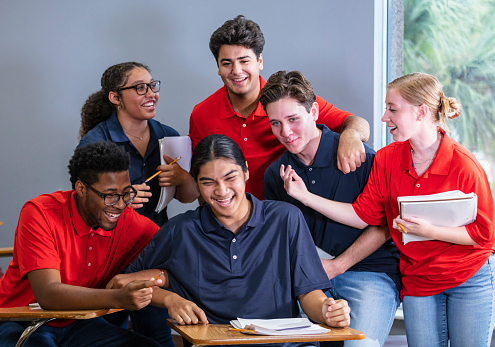 A multiracial group of six high school students sitting together in a classroom. The students are in two teams, wearing either blue or red shirts. They could be participating in an after school activity, perhaps a debate team or math club.