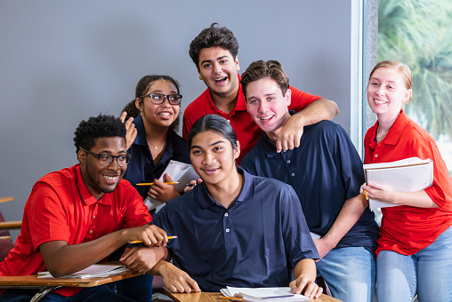 A multiracial group of six high school students sitting together in a classroom. The students are in two teams, wearing either blue or red shirts. They could be participating in an after school activity, perhaps a debate team or math club.