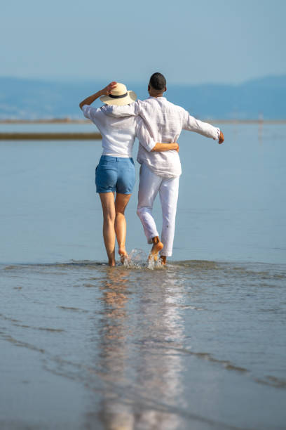 couple walking on beach - women wading sun hat summer foto e immagini stock