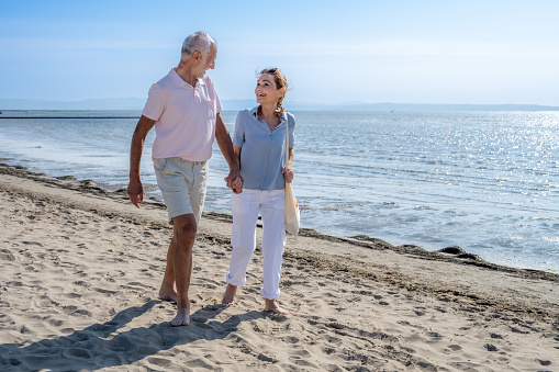 Smiling senior couple with holding hands walking on beach against sky during vacation in sunny day.