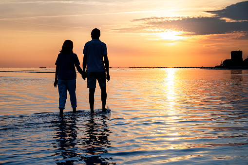 Rear view of senior couple with holding hands walking on beach while vacation during sunset.