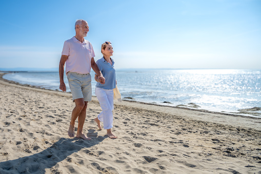 Smiling senior couple with holding hands walking on beach against sky during vacation in sunny day.