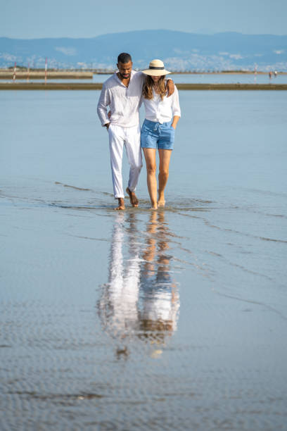 couple walking on beach - women wading sun hat summer foto e immagini stock