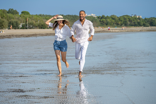 Front view of smiling couple running while holding hands on beach during vacation.