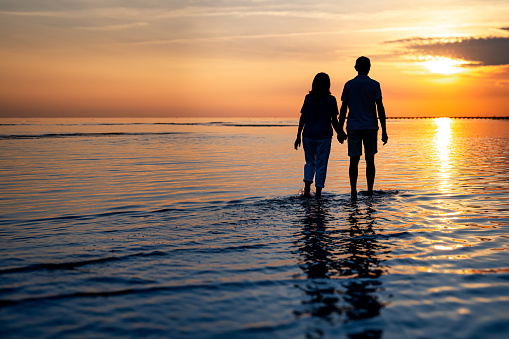 Rear view of senior couple with holding hands walking on beach while vacation during sunset.
