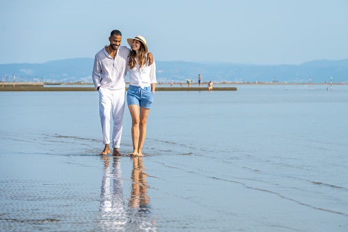 Front view of couple with arm around walking on beach during vacation in sunny day.