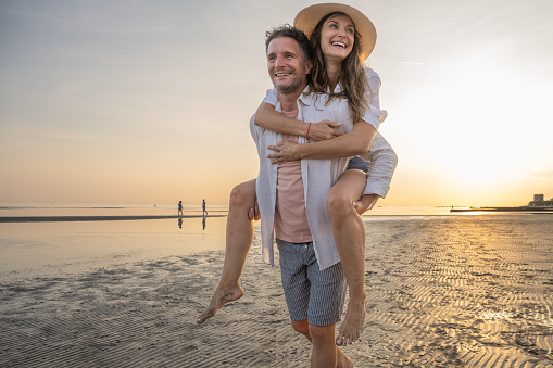 Mature man carrying woman piggyback while walking on beach during vacation in sunny day.