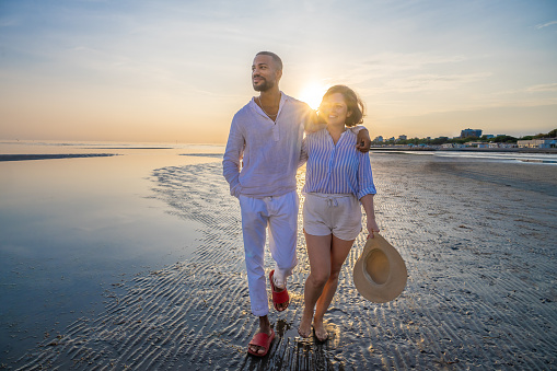 Smiling couple with arm around walking on beach during vacation in sunny day.