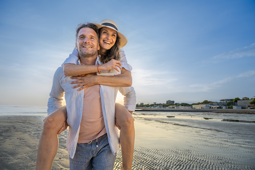 Mature man carrying woman piggyback while walking on beach during vacation in sunny day.