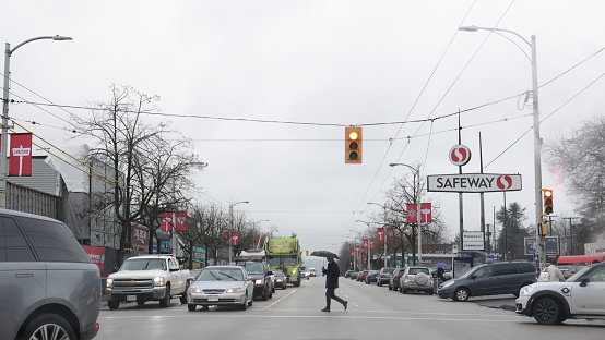 Vancouver, Canada - December 21, 2023: Looking west at traffic on the 2300-block of West 4th Avenue. A pedestrian holding an umbrella crosses West 4th at Vine Street. Winter morning in the Kitsilano neighbourhood on the Westside.