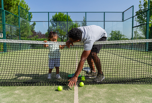 Full body African American dad carrying little kid and taking ball from ground while bending over net during paddle tennis training on court on sunny summer day