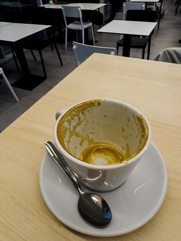 the solitude of a cafe table after a patron has departed, leaving behind a cup stained with the remnants of a pumpkin spice latte. The starkness of the empty chairs and tables in the background contrasts with the detailed texture of the coffee residue, suggesting the quiet after the morning rush. The spoon, resting on the saucer, indicates the end of a moment of enjoyment, and the wooden table provides a warm, inviting texture, echoing the comfort of the cafe