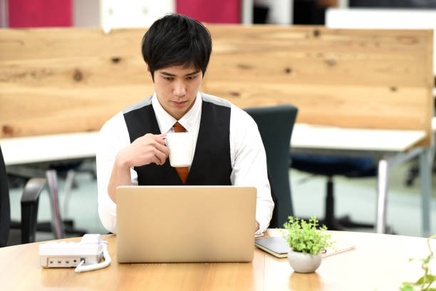 istock Asian young male office worker working on laptop while having a drink indoors 1907520562