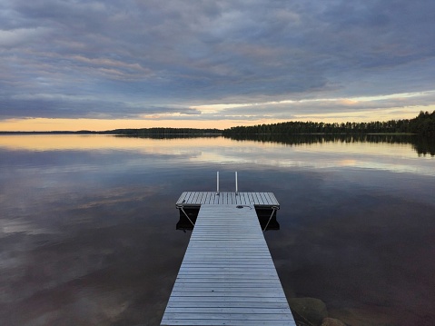 Heavy clouds hanging and mirroring over calm Lake Saimaa surface. Thin gap of bright yellow sunset shines in the horizon. Small pier in the middle of photo.