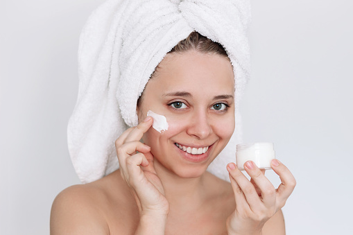 Young caucasian smiling woman with towel on her head after shower touching face with hand applying a smear of cream on the skin isolated on white background. Moisturizing cream. Cosmetology and beauty