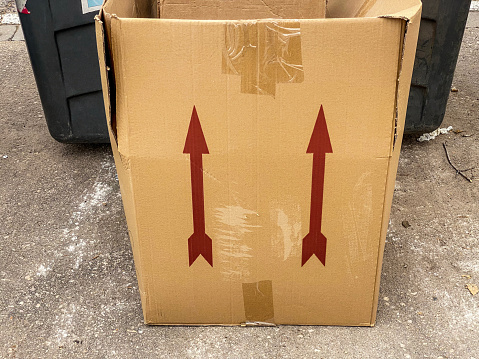 High angle close-up view of a cardboard box lying on the sidewalk with two arrow symbols pointing up printed on it