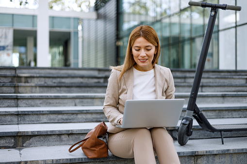 A businesswoman combines green commuting with connectivity, working on her laptop while sitting on a staircase next to her electric scooter in the city