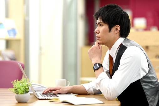 Young Asian male office worker thinking while working on laptop in office