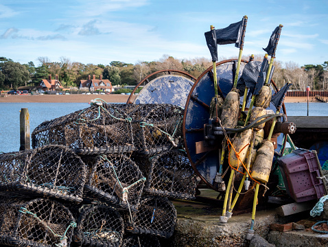 A pile of crab and lobster pots stacked on the riverbank of the River Deben at Felixstowe Ferry in Suffolk, Eastern England, together with flags, marker buoys and various other items relating to the crustacean fishing industry. The tiny hamlet of Bawdsey is across the estuary.