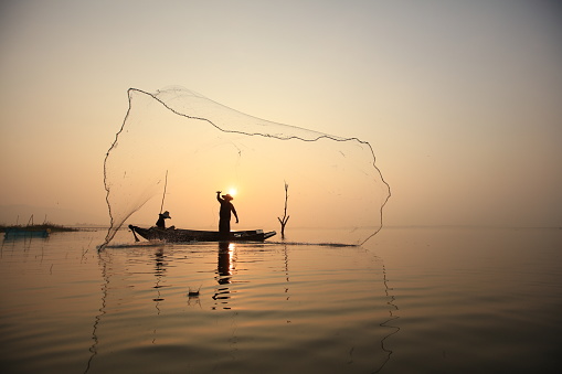Silhouette fisherman on boat throwing net in lake