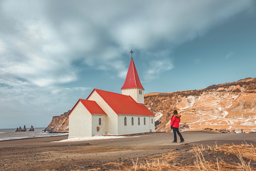 Tourist woman in red jacket enjoy looking view of Reyniskirkja church among the mountain near the beach on winter in Vik town at South of Iceland