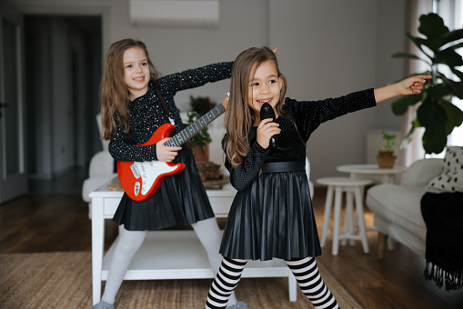 Funny adorable happy kid girls player guitars and sing in microphone. Two happy beautiful caucasian laughing little girls playing guitar and applauding at home