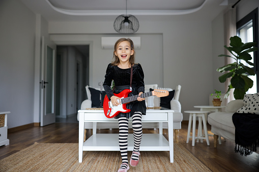 Confined 6 years old little girl learning electric guitar, wearing and princess dress. Young girl rocking in her girlish room on an elecric guitar. Musician of the future. Girl rockstar.